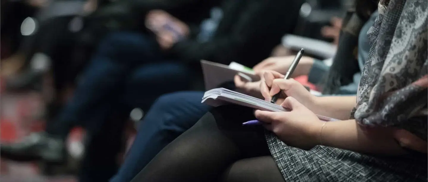 Woman sitting at a conference, writing notes in her notebook with a pen