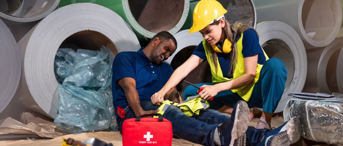 Woman in a hi-vis jacket and a helmet tying a bandage around the leg of a man lying on the ground